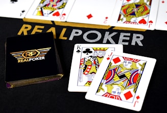 Poker-themed setup featuring a deck of cards on a table with the words 'REAL POKER' visible. Two cards, the King of Clubs and Jack of Diamonds, are prominently displayed. The design is vibrant, with detailed illustrations on the cards.