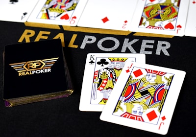 Poker-themed setup featuring a deck of cards on a table with the words 'REAL POKER' visible. Two cards, the King of Clubs and Jack of Diamonds, are prominently displayed. The design is vibrant, with detailed illustrations on the cards.