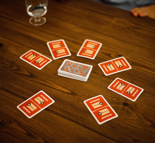 A wooden table with several playing cards arranged in a circle around a stack of cards in the center. Each card has a red back with text and illustrations. Two drinking glasses, one with a clear liquid, are also on the table, with a person's arm visible in the background.