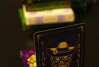 A playing card featuring a stylized sugar skull design, labeled 'Joker', is prominently displayed in the foreground. The card has an intricate design with a hat on the skull and decorative elements in gold. In the background, multiple stacks of board games or boxes are slightly out of focus, placed on a dark tabletop.