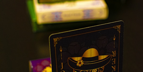 A playing card featuring a stylized sugar skull design, labeled 'Joker', is prominently displayed in the foreground. The card has an intricate design with a hat on the skull and decorative elements in gold. In the background, multiple stacks of board games or boxes are slightly out of focus, placed on a dark tabletop.