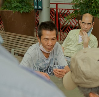Several individuals gather outdoors, sitting on benches and engaging in a game of cards. The focal point is on a man holding a fan of playing cards, with others observing or participating. Potted plants and a red railing are visible in the background, contributing to the setting's casual and social atmosphere.