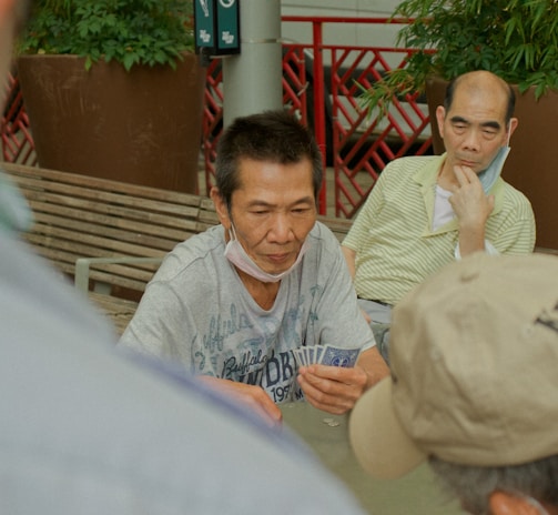 Several individuals gather outdoors, sitting on benches and engaging in a game of cards. The focal point is on a man holding a fan of playing cards, with others observing or participating. Potted plants and a red railing are visible in the background, contributing to the setting's casual and social atmosphere.