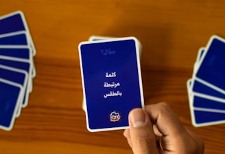 A hand holds a playing card with Arabic text printed on it. Several other cards are spread out on a wooden surface in different positions.