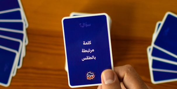 A hand holds a playing card with Arabic text printed on it. Several other cards are spread out on a wooden surface in different positions.