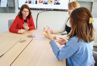 Three people are sitting around a light wooden table playing a card game. One person in a red top holds a hand of cards, while the others are also engaged with their cards. A large screen on the wall displays a website with the hashtag #LoveWallsio.