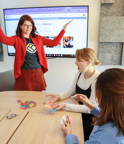 Four people are engaged in a lively game around a wooden table. One person stands with raised arms, while three others are seated, focusing on colorful cards in their hands. There is a computer monitor in the background displaying a webpage, and the room features a modern design with gray acoustic panels on the walls.