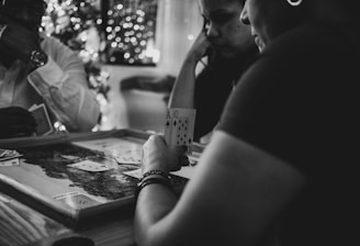 Three people are engaged in a card game, sitting around a table in a cozy, dimly lit setting. One player is holding an Ace and a Ten of diamonds. There are fairy lights and a blurred Christmas tree in the background, adding a festive and warm atmosphere to the scene.