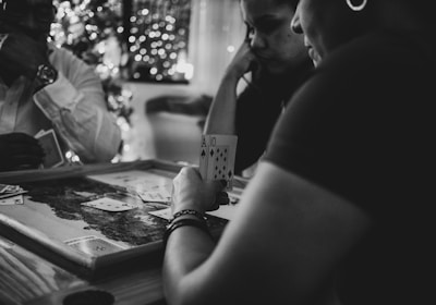 Three people are engaged in a card game, sitting around a table in a cozy, dimly lit setting. One player is holding an Ace and a Ten of diamonds. There are fairy lights and a blurred Christmas tree in the background, adding a festive and warm atmosphere to the scene.